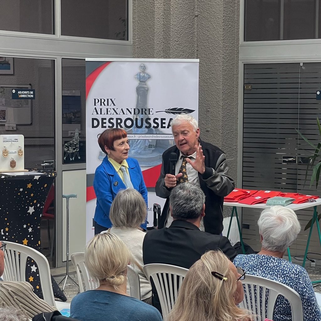 Older man speaks into a microphone and gestures as he addresses an audience, next to a woman in a blue jacket at an award event poster reading 'Prix Alexandre Desrousseau' behind them.